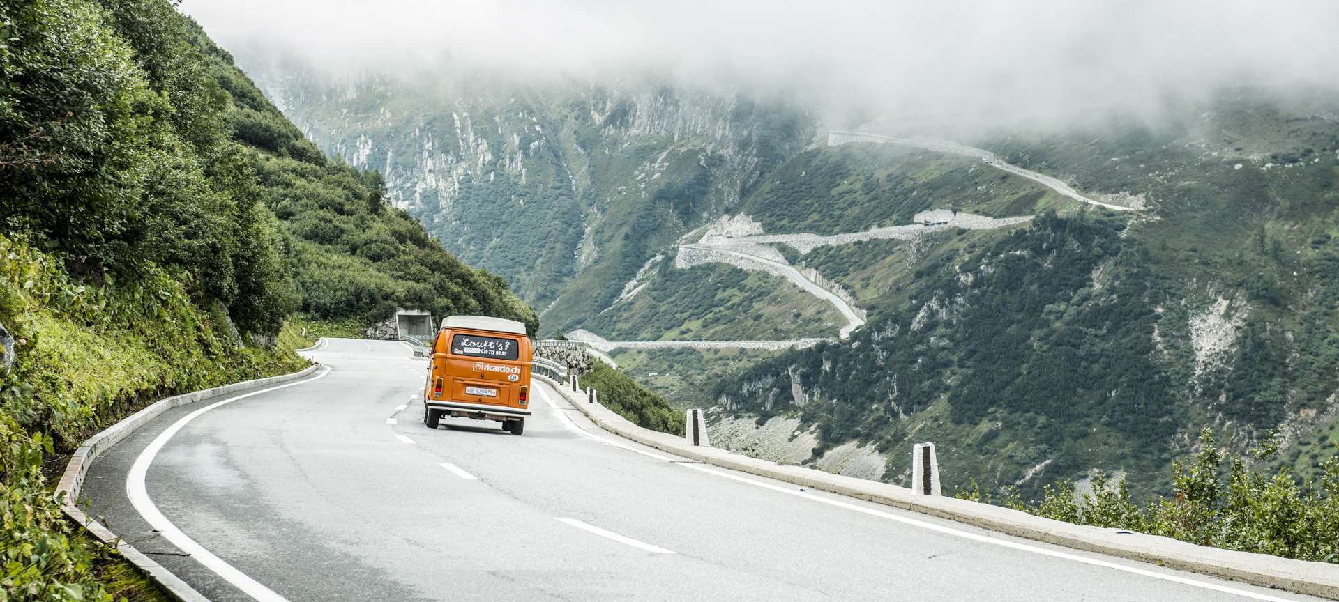 Passage de col, bus VW de location avec inscriptions à l'arrière dans montagnes suisses